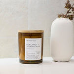 A candle in an amber glass jar with a light wood lid and a white label reading 'Bergamot + Jasmine Musk' by Re:Union label, styled on a white surface next to a white ceramic vase with dried branches against a light textured wall.