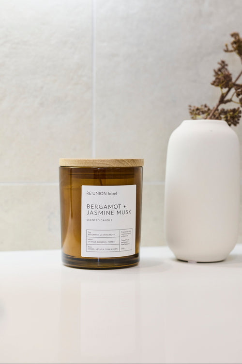 A candle in an amber glass jar with a light wood lid and a white label reading 'Bergamot + Jasmine Musk' by Re:Union label, styled on a white surface next to a white ceramic vase with dried branches against a light textured wall.