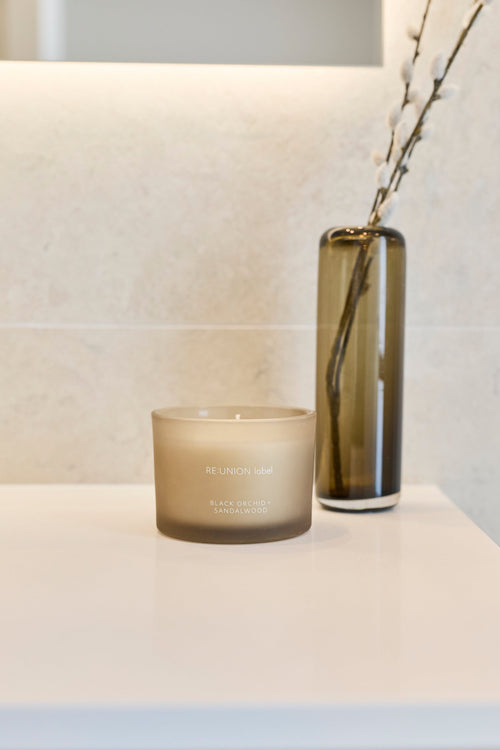 A frosted glass jar candle sits on a white countertop beside a tall smoky amber glass vase holding dried branches, with a light stone tile backsplash behind.