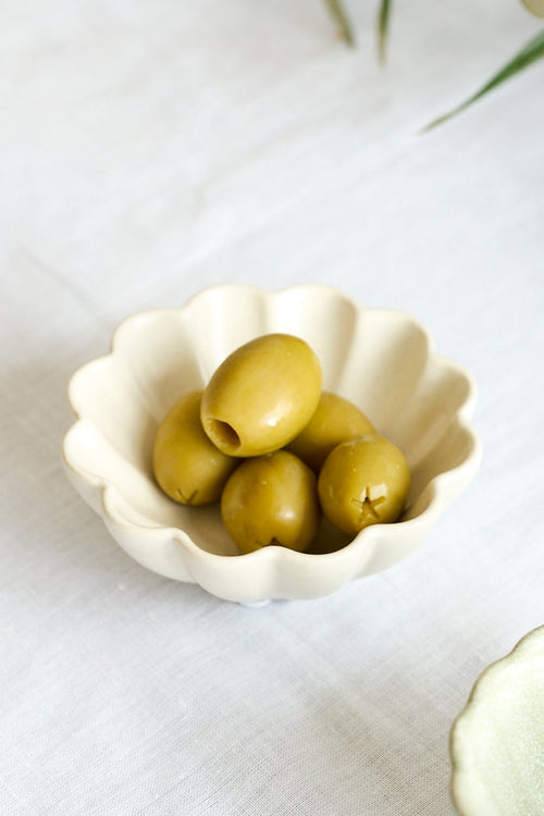 Two small scalloped ceramic bowls on a white fabric surface, with one bowl containing green olives and the other empty, styled with olive branches in the background.