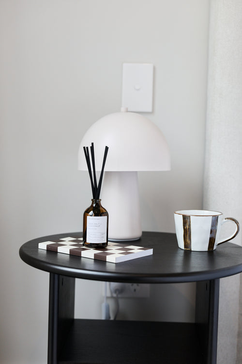 A modern white mushroom-shaped table lamp on a black round side table, styled with a reed diffuser, a checkered book, and a white mug with gold trim against a light gray wall.