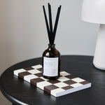 An amber glass reed diffuser bottle with black reeds stands on a black-and-white checkerboard tray on a round black table, with a white lamp and a white cup with gold trim in the background.