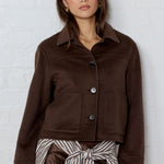 Woman wearing a brown jacket and striped skirt against a white brick wall.
