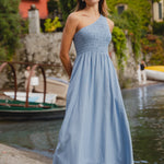 Woman in a blue dress standing by a waterfront with boats and greenery in the background