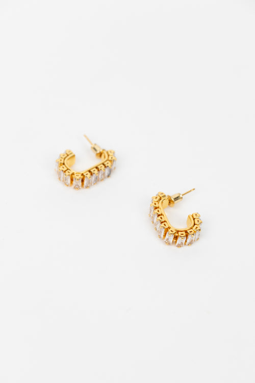 A pair of gold crescent-shaped hoop earrings adorned with clear baguette crystals, displayed on a clean white background.