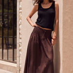 Woman in black tank top and brown wide-leg pants standing against a beige wall.