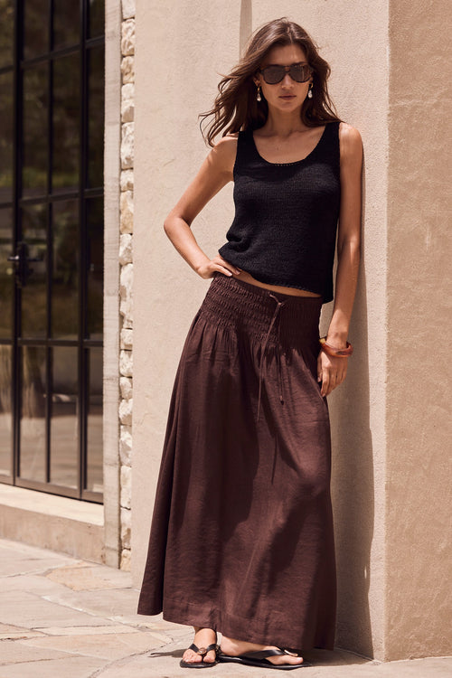 Woman in black tank top and brown wide-leg pants standing against a beige wall.