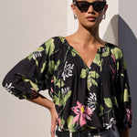 A woman modeling a black floral blouse with green and pink botanical print, paired with white shorts, standing against a light wall with strong sunlight and shadows.