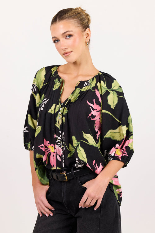 A woman modeling a black blouse with a vibrant green, pink, and white tropical floral print, paired with black jeans, against a plain white studio background.