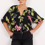 A woman modeling a black short-sleeve blouse with a vibrant green, pink, and white floral print, paired with black jeans and sunglasses, set against a plain white studio background.