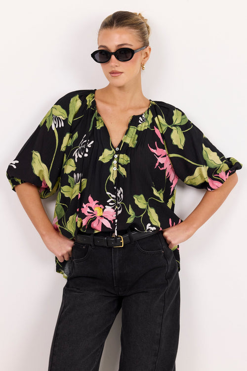 A woman modeling a black short-sleeve blouse with a vibrant green, pink, and white floral print, paired with black jeans and sunglasses, set against a plain white studio background.