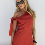 A woman modeling a terracotta red one-shoulder linen dress with a tie detail on the shoulder, styled with black sunglasses against a white wall.