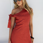 A woman modeling a cinnamon red one-shoulder linen dress with a side tie detail, paired with black sunglasses, standing against a white wall.