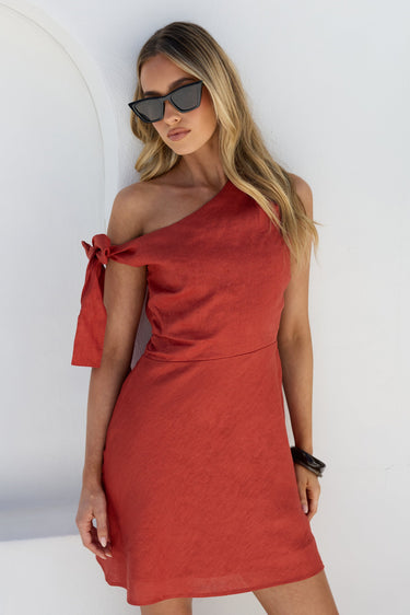 A woman modeling a cinnamon red one-shoulder linen dress with a side tie detail, paired with black sunglasses, standing against a white wall.