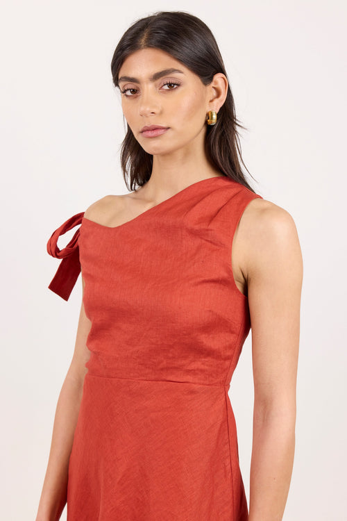 A woman modeling a rust-orange one-shoulder dress with a tie detail on the shoulder, photographed against a clean off-white studio background.