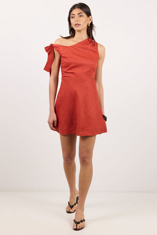 A woman modeling a terracotta orange-red off-the-shoulder mini dress with a ruffled sleeve detail, paired with black sandals, against a plain white studio background.