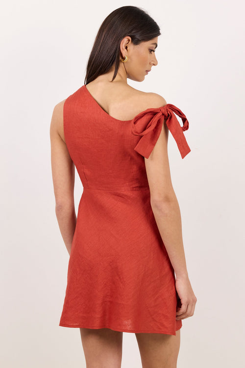 A woman modeling a rust-orange one-shoulder dress with a large bow detail on the shoulder, shown from the back against a plain light background.