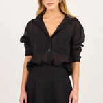 A woman modeling a black sheer long-sleeve blouse with puffed shoulders and a black mini skirt, standing against a plain off-white studio background.