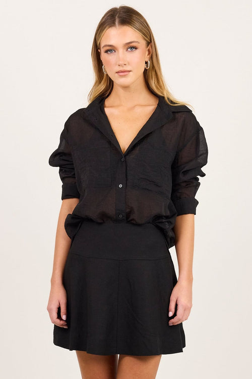 A woman modeling a black sheer long-sleeve blouse with puffed shoulders and a black mini skirt, standing against a plain off-white studio background.