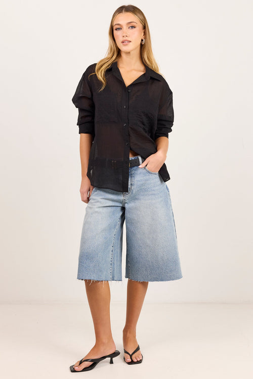 A woman modeling a black sheer long-sleeve button-up shirt paired with light-wash denim culotte shorts and black heeled sandals, standing against a plain off-white studio background.