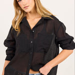 A woman wearing a black sheer button-up shirt with textured side panels, paired with light-wash jeans and a black belt, against a plain off-white background.