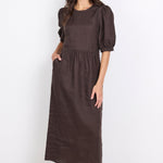 A woman modeling a dark brown midi dress with puffed short sleeves, a round neckline, and side pockets, standing against a plain white studio background.