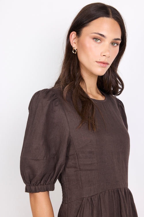 A woman wearing a dark brown linen dress with puffed elbow-length sleeves and a gathered waist, photographed against a plain white studio background.