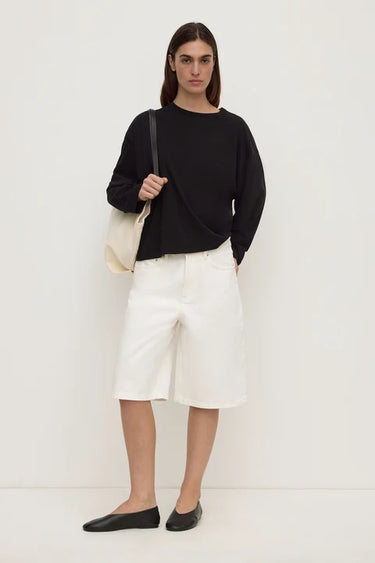 A woman stands against a plain off-white studio wall wearing a black long-sleeve top, white knee-length shorts, and black flats, while carrying a cream shoulder bag.