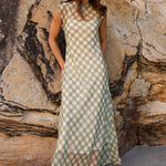 A woman wearing a sleeveless olive green and beige gingham maxi dress stands in front of a large rock wall under a wooden structure.