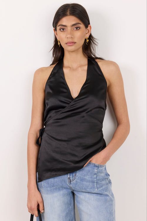 A woman modeling a black satin halter wrap top with light blue jeans, standing against a plain off-white background.