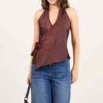 A woman modeling a sleeveless brown wrap-style top with side ties, paired with wide-leg blue jeans, black sandals, and a small brown handbag, standing against a plain light studio background.