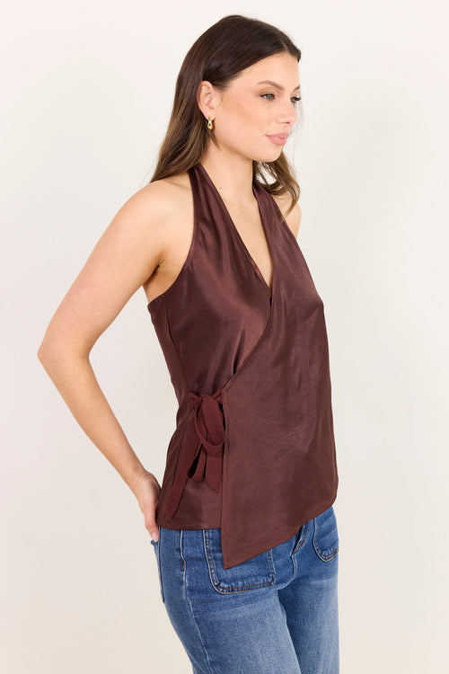 A woman wearing a sleeveless halter-neck wrap top in a deep brownish-rose color, paired with blue jeans, standing against a plain off-white background.
