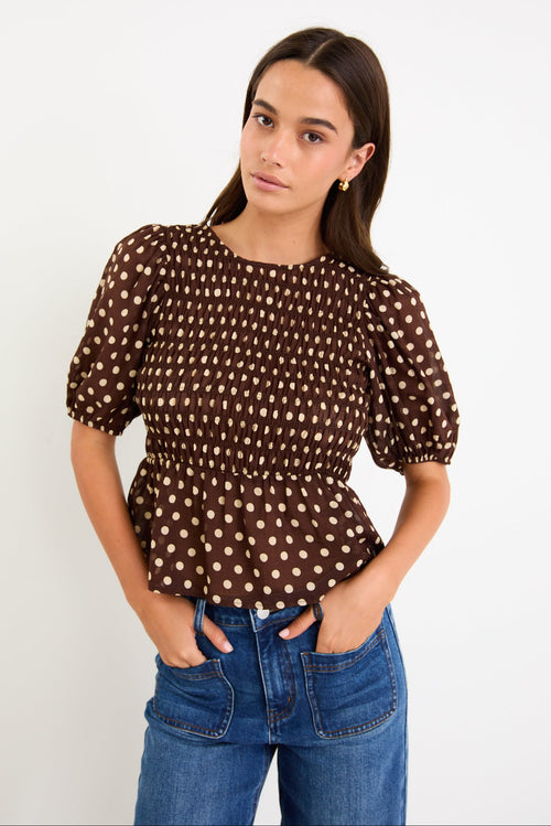 Model wearing a chocolate brown polka dot short sleeve top with a shirred bodice and peplum hem
