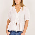 A woman modeling a white short-sleeve blouse with a V-neck, tie-front detail, and peplum hem, paired with blue jeans, against a plain off-white studio background.
