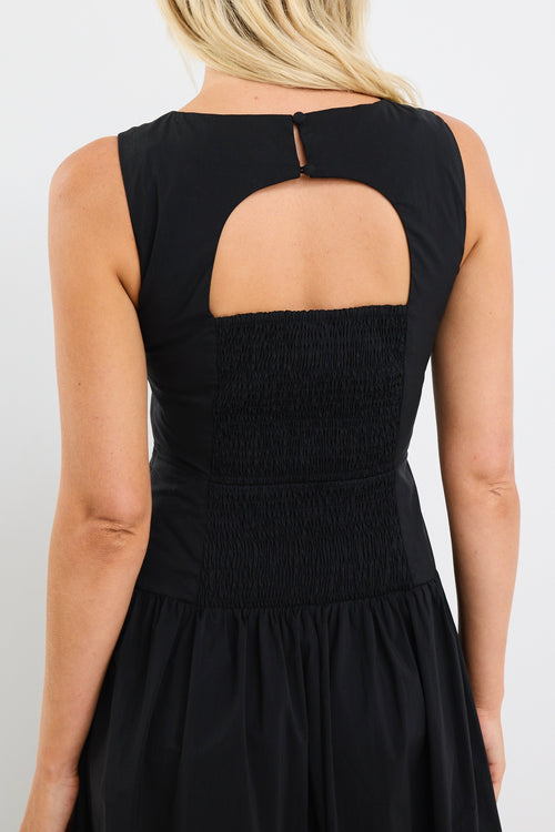 Woman wearing a sleeveless black midi dress with a fitted bodice and gathered skirt, styled with black braided sandals and gold hoop earrings.







