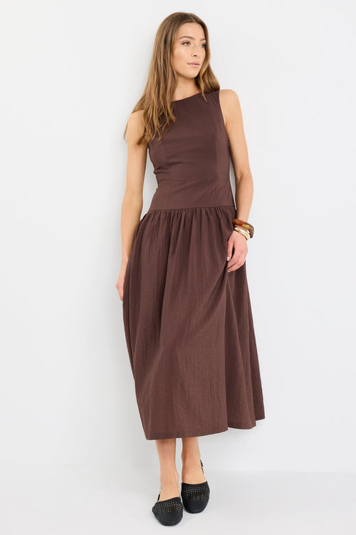 Woman wearing a chocolate brown sleeveless maxi dress with a structured bodice and flowing skirt, styled with black flat shoes