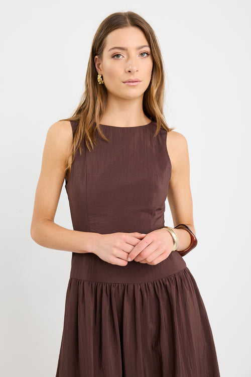 Woman wearing a chocolate brown sleeveless maxi dress with a structured bodice and flowing skirt, styled with black flat shoes