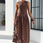 A woman wearing a sleeveless brown midi dress with a white polka dot pattern, styled with sunglasses, a small handbag, and sandals, walking outdoors in front of a white building with a large black-framed window.