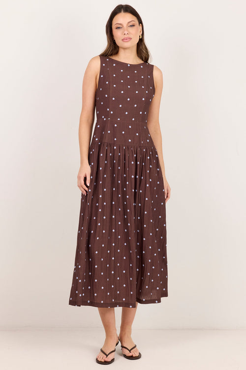 A woman modeling a sleeveless brown midi dress with a white polka dot pattern, standing against a plain off-white studio background.