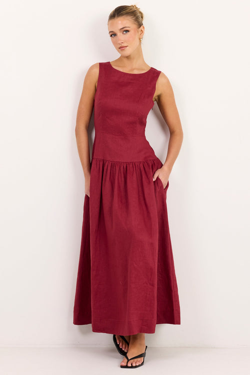 A woman modeling a sleeveless, floor-length maroon dress with side pockets, standing against a plain white studio background.
