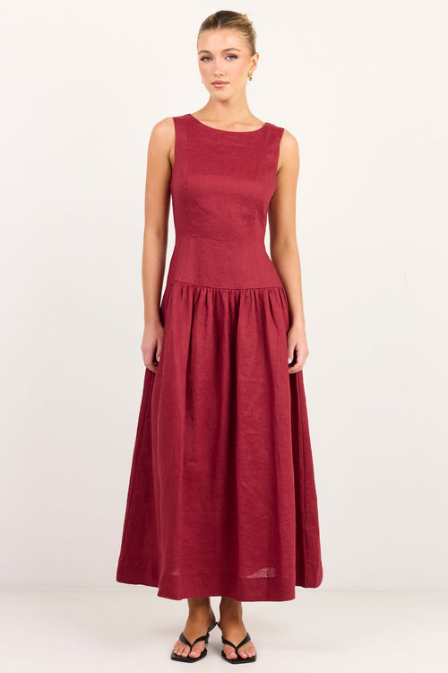 A woman modeling a sleeveless, high-neck, midi-length A-line dress in a deep burgundy or maroon shade, standing against a plain white studio background.