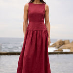 A woman stands outdoors by the water wearing a sleeveless, high-waisted maxi dress in a deep burgundy red. The dress has a structured bodice and a flowing skirt, paired with brown sandals.