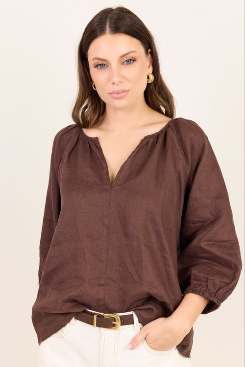 A woman wearing a brown long-sleeve linen blouse with a split neckline and gathered cuffs, paired with white pants and a brown belt, against a plain off-white background.