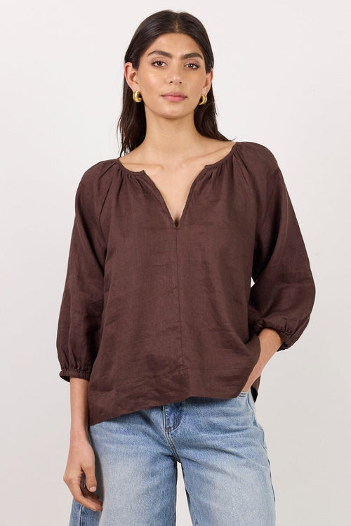 A woman modeling a dark brown long-sleeve blouse with a split neckline and light-wash denim cutoff shorts against a plain white studio background.