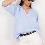 A woman wearing a light blue linen blouse, white trousers, and black sunglasses, posing against a plain white wall.