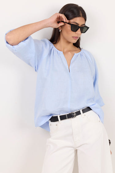 A woman wearing a light blue linen blouse, white trousers, and black sunglasses, posing against a plain white wall.