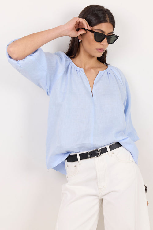 A woman wearing a light blue linen blouse, white trousers, and black sunglasses, posing against a plain white wall.