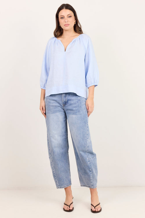 A woman modeling a light blue blouse with three-quarter sleeves and a split neckline, paired with light-wash cropped jeans and black sandals, against a plain off-white studio background.
