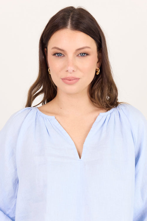 A woman wearing a light blue, loose-fitting blouse with voluminous puff sleeves and a split V-neckline, photographed against a solid off-white background.