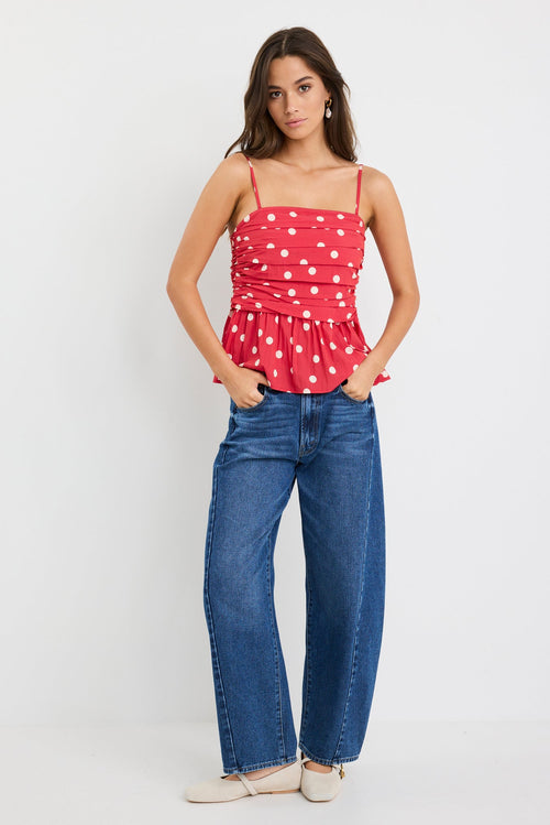 Woman wearing a red polka dot ruched cami top with thin straps and a peplum hem, styled with blue jeans against a plain white background.